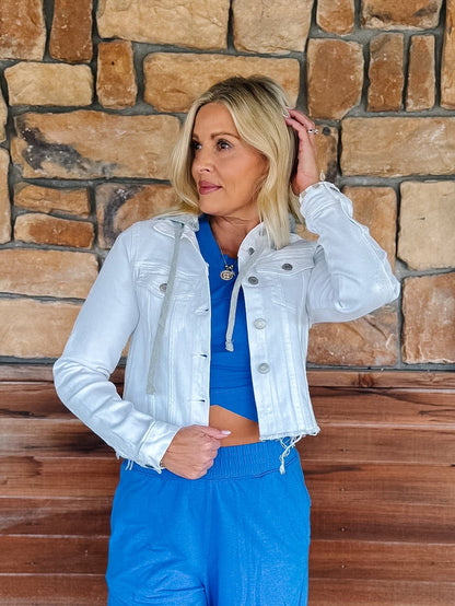 A woman posing in front of a rustic stone wall, wearing a light silver-white cropped denim jacket over a bright blue crop top and matching blue sweatpants.