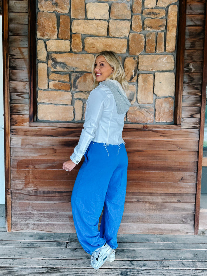 A woman posing in a light blue hooded sweatshirt and bright blue sweatpants against a rustic wooden and stone wall background.