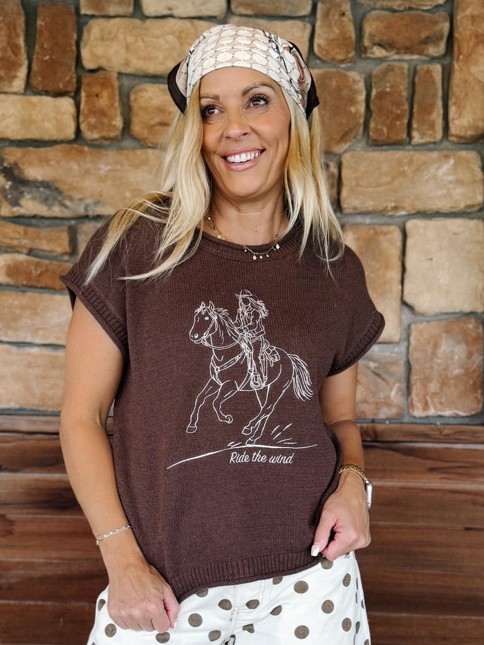 A smiling blonde woman wearing a brown short-sleeve knit t-shirt with a white line art graphic of a cowboy riding a horse and the text 'Ride the wind'. She is also wearing a cream patterned headband and white pants with brown polka dots, standing in front of a rustic stone wall.