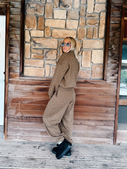 A woman wearing a matching brown polka-dot lounge set stands on a rustic wooden porch in front of a stone wall.