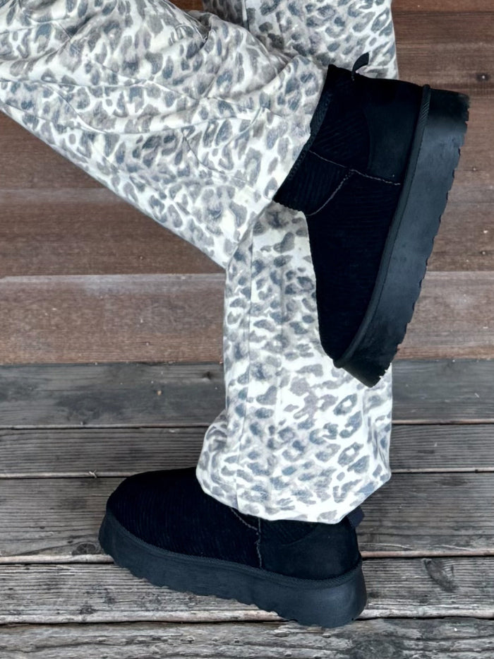 Black suede Chelsea-style slipper boots with chunky black soles, worn with white and gray leopard print pants on a weathered wooden deck.